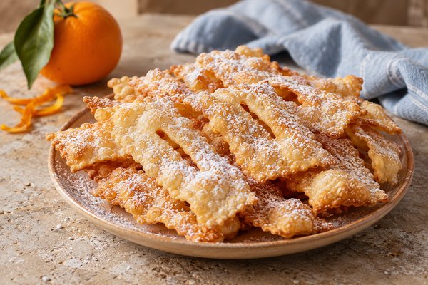 Oreillettes fines dorées et saupoudrées de sucre glace dans une assiette, avec une orange